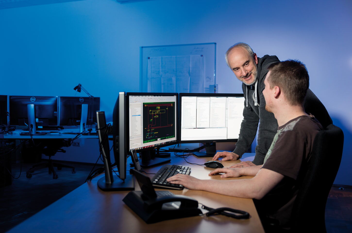 Ausbilder gibt jungen Mitarbeiter Tipps vor 3 Computerbildschirmen